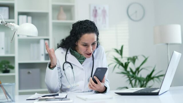 Happy excited female doctor read great news on smartphone while sitting at desk at workplace in modern hospital clinic. Joyful surprised brunette medical worker physician celebrates success in office
