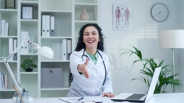 Smiling positive female doctor in white coat waving hand looking at camera and giving hand for greeting at desk at workplace in modern hospital clinic. Friendly brunette woman medical worker physician