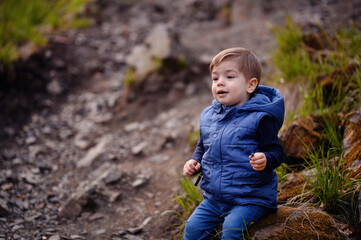 Little toddler boy in blue spring warm outfit sitting on stone in forest
