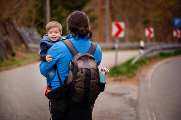 Man from back in glasses with long dark hair and photo backpack holding little toddler boy on arms
