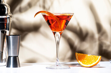 Martinez orange cocktail drink with red vermouth, liqueur, bitter, citrus zest and ice in martini glass. Light beige background, hard light, shadow pattern