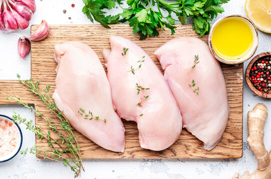 Raw chicken fillet on rustic wooden cutting board prepared for cooking with garlic, thyme, spices and pepper. White kitchen table, top view