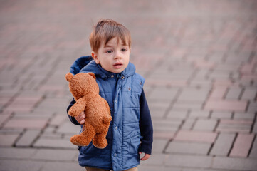 Little toddler boy in blue spring-autumn outfit looking forward holding teddy bear toy on street
