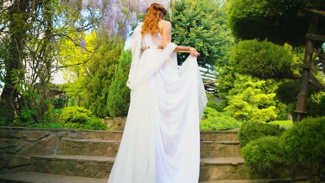 Fantasy woman greek goddess walks in spring garden purple flowers green tree nature day sun light. Sexy girl queen antique style white silk dress gold diadem on red hair, lady bride back rear view 4k
