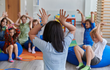 A happy female teacher sitting and playing hand games with a group of little schoolchildren