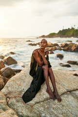 Fototapeta na wymiar Androgynous ethnic fashion model in luxury dress, jewelry sits on rocks by ocean. Gay black person in jewellery, posh clothes poses gracefully in tropical seaside location. Pride LGBTQ bipoc concept.