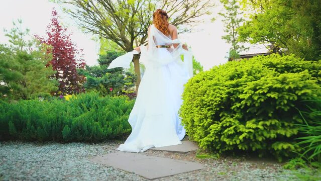 Fantasy happy woman greek goddess walks in spring garden fresh green tree nature day sun light sky. Sexy girl queen antique style white silk dress hem fly in wind. red hair lady bride back rear view