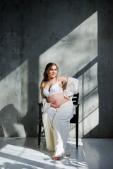 A stylish young pregnant woman in a white suit is sitting on a chair on a gray background. Pregnant woman in white underwear.