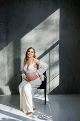 A stylish young pregnant woman in a white suit is sitting on a chair on a gray background. Pregnant woman in white underwear.