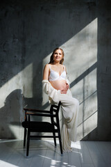 A stylish young pregnant woman in a white suit is sitting on a chair on a gray background. Pregnant woman in white underwear.