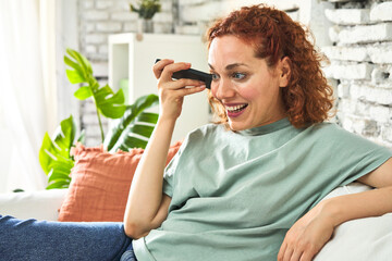 Happy young redhead listening to voice message on phone at home