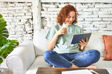 .Redhead woman on sofa, sipping milk, watching videos on tablet for a cozy morning.