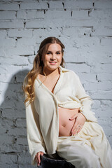 A stylish pregnant young woman in a white suit sits against a white brick wall and touches her belly with a gentle smile. Awaiting birth. Care and motherhood. Love and tenderness.