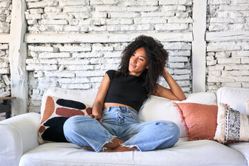 Afro-haired woman relaxes on sofa, basking in soft morning light at home.