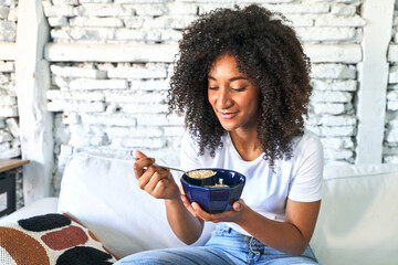 African-American woman savors a comforting bowl of oatmeal on the sofa, starting her day with...