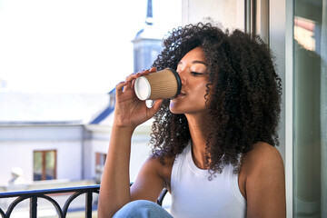 African-American woman peacefully sips a takeaway coffee on her balcony, embracing the serene cityscape.