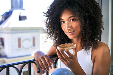 African-American woman peacefully enjoys a rice cake with peanut butter on her balcony, embracing...