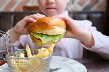 Cute healthy preschool girl eats a hamburger sitting in a cafe. A happy child eats junk food in a...
