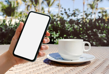 Hand with mobile phone and white coffee cup on table in outdoor cafe.