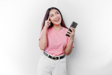 A thoughtful young woman wearing pink t-shirt, holding her chin and her phone isolated by white background