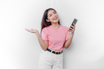 A portrait of a happy Asian woman wearing pink t-shirt, holding her phone, and pointing copy space on beside her, isolated by white background