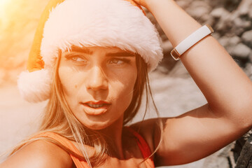 Woman summer travel sea. Happy tourist in red bikini and Santas hat enjoy taking picture outdoors for memories. Woman traveler posing on the beach surrounded by volcanic mountains, sharing travel joy