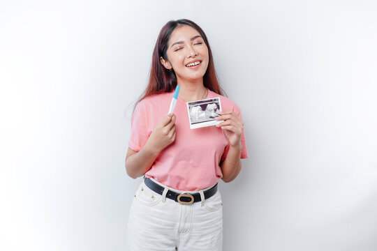 Happy young woman wearing pink t-shirt showing her pregnancy test and ultrasound picture, isolated on white background, pregnancy concept