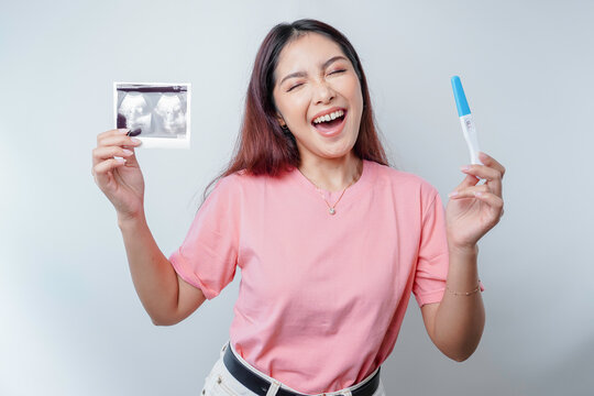 Happy and proud young woman wearing pink t-shirt showing her pregnancy test and ultrasound picture, isolated on white background, pregnancy concept