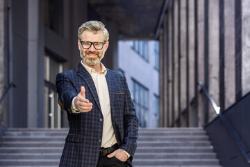 Mature successful gray-haired businessman smiling and looking at camera, holding hand in front handshake greeting gesture, experienced boss.