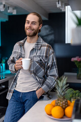 Man with beard holding cup with coffee, standing by kitchen counter with modern light interior.