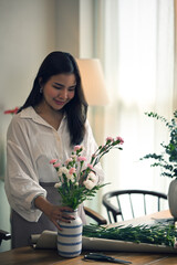 A gorgeous young Asian woman enjoys arranging a vase with beautiful flowers at home.