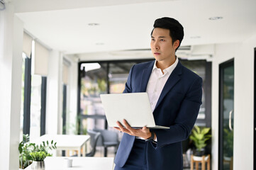 Smart and focused Asian businessman stands in his office with his laptop computer.