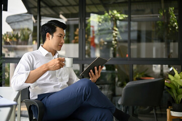 An Asian businessman focuses on reading a book while sipping his morning coffee