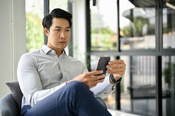 Handsome Asian businessman using his smartphone while relaxing in an office lounge.