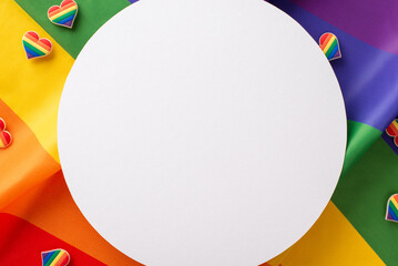 A top view flat lay of LGBT-themed parade accessories, featuring pin badges on a waving rainbow flag as the backdrop, empty circle for text or message