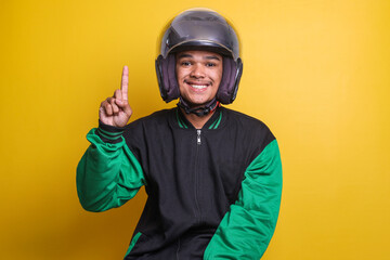 Asian online taxi driver wearing green jacket and helmet pointing to empty space with finger isolated over yellow background