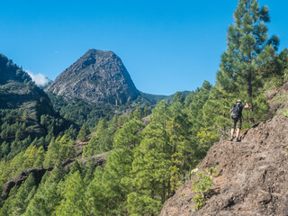 Man hiker climbing at steep hiking trail to La Laja with volcanic rock formation Roque de Ojila in...