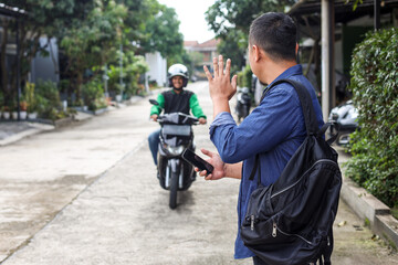 Man standing in front of a house ordering motorcycle taxi by waving his hand 
