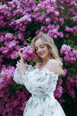 Beautiful, stylish, cute blonde in a white dress with a green pattern as a model for a photo shoot in lilac. They made luxurious portraits of her in the green garden of the park.