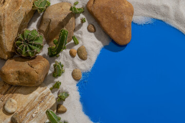 Against a sandy background, several stones in different sizes are arranged with many green Cacti beside a lake with blue water
