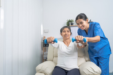 Fototapeta premium Young pretty asian physiotherapist helping senior female patient holding dumbbell in physical therapy session, concept of World Confederation for Physical Therapy.