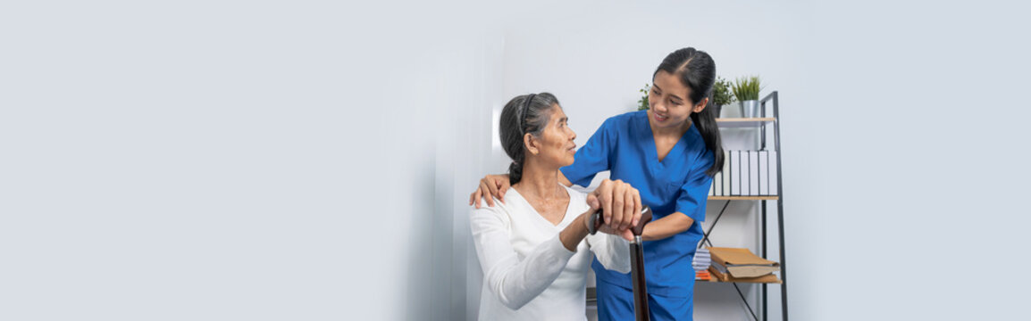 Attentive practitioner nurse assisting physical therapy elderly woman on a walking wood standard cane in disability nursing rehabilitation center, physical therapy encourage hands.