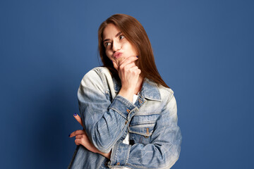 Portrait of funny, pensive girl wearing casual style clothes grimacing with doubtful face over blue color studio background