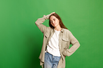 Portrait with one doubtful young girl, teenager in casual clothes touching head and looking away with pensive face over green color studio background