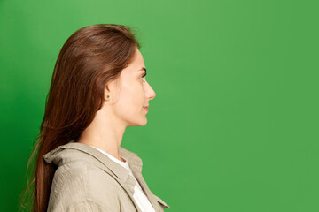 Fototapeta premium Side view portrait of young pretty, positive girl, student with big eyes posing with calm face over green color studio background
