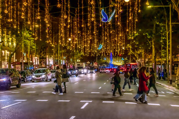 Fototapeta premium Barcelona, Spain - November 26,2021: Blue glowing butterflies and golden garlands hang over the road on Passeig de Gracia in Barcelona at night. Cars and people move under festive illumination at dusk