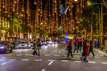 Zelfklevend Fotobehang Barcelona, Spain - November 26,2021: Blue glowing butterflies and golden garlands hang over the road on Passeig de Gracia in Barcelona at night. Cars and people move under festive illumination at dusk © ioanna_alexa