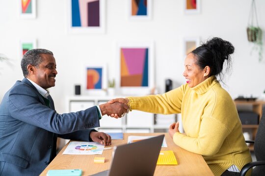 Side view of smiling Ethiopian woman and man in smart casual clothes shaking hands after successful discussion while looking at each other in modern office