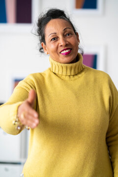 Positive ethnic mature woman with makeup in yellow sweater holding out hand for handshake while looking at camera