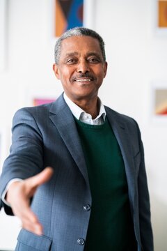 Positive successful mature black entrepreneur in suit smiling while stretching hand at camera in greeting gesture